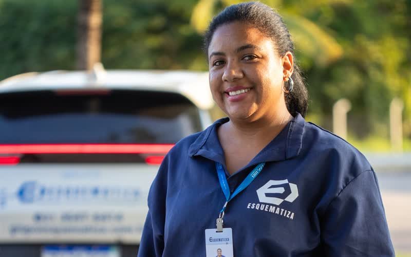 Auxiliar de serviços gerais da Esquematiza uniformizada, sorrindo, diante de uma viatura da empresa.