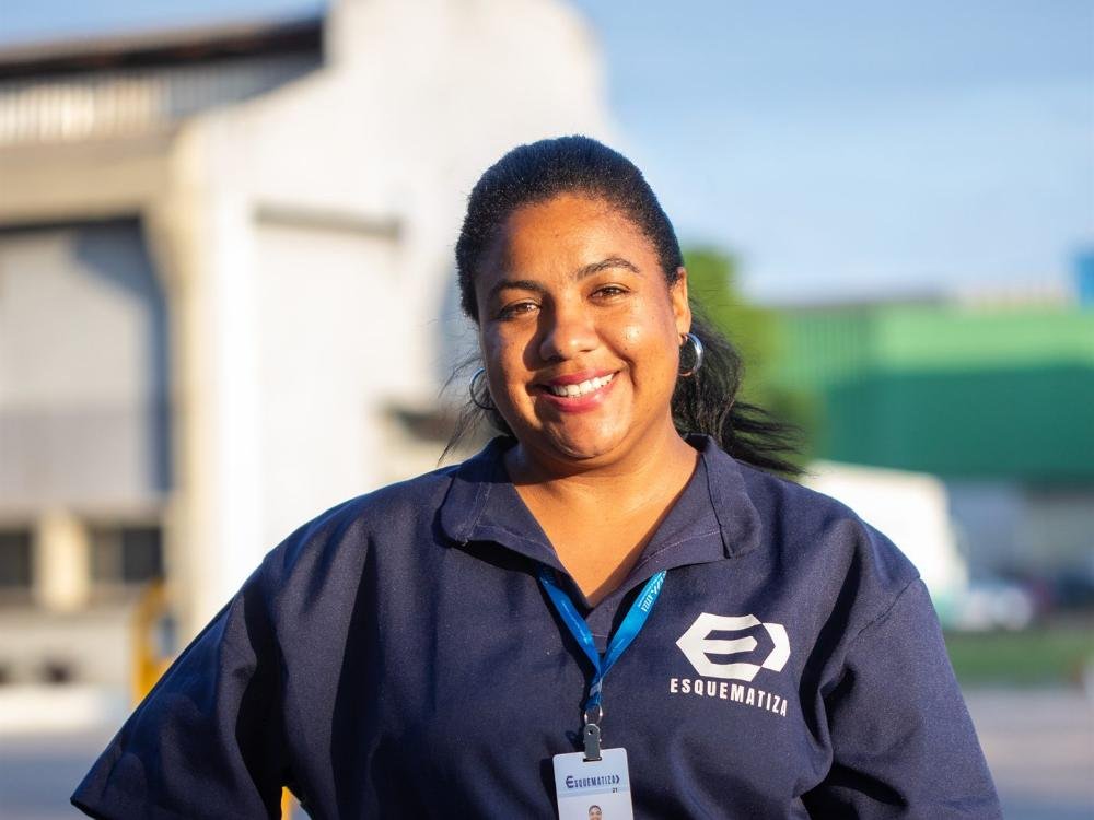 Auxiliar de serviços gerais da Esquematiza, uniformizada e com crachá, sorrindo em frente a um galpão industrial.